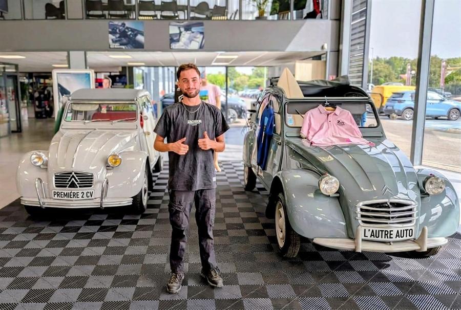 Jeune en alternance au GEIQ AC2R SUD, pouces levés devant deux Citroën 2CV dans le hall d’une concession